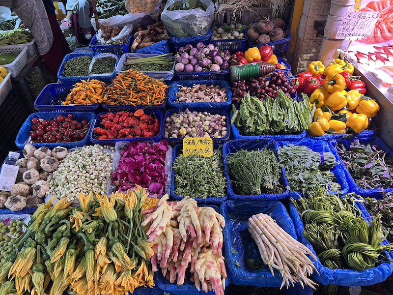 地州山茅野菜专卖店售卖的菜蔬品种繁多，照片中可见草芽、海菜花、南瓜花、臭菜（羽叶金合欢）、夜来香花蕾、玫瑰花瓣、茉莉花、百合、四棱豆、新鲜的涮涮辣……  摄影/半夏